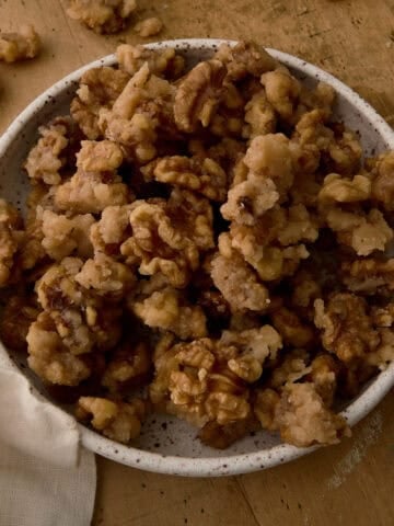 A bowl of candied walnuts is surrounded by random walnuts.