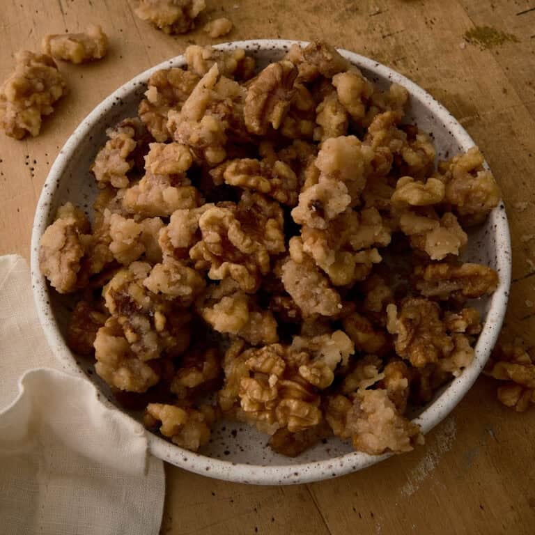 A bowl of candied walnuts is surrounded by random walnuts.