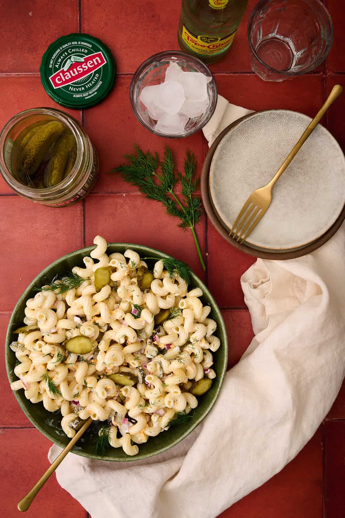 An open jar of pickles, a plate, and the bowl of dill pickle pasta salad are surrounded by a white napkin.