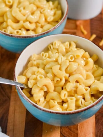 Two bowls of instant pot mac and cheese are on a wooden cutting board with shredded cheese around the bowls.