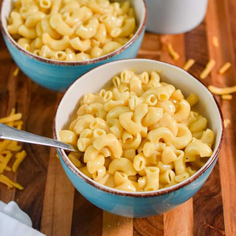 Two bowls of instant pot mac and cheese are on a wooden cutting board with shredded cheese around the bowls.