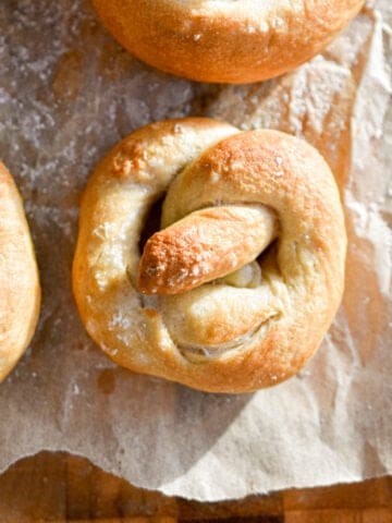 A sourdough pretzel is next to some other pretzels on a piece of parchment paper on top of a cutting board.