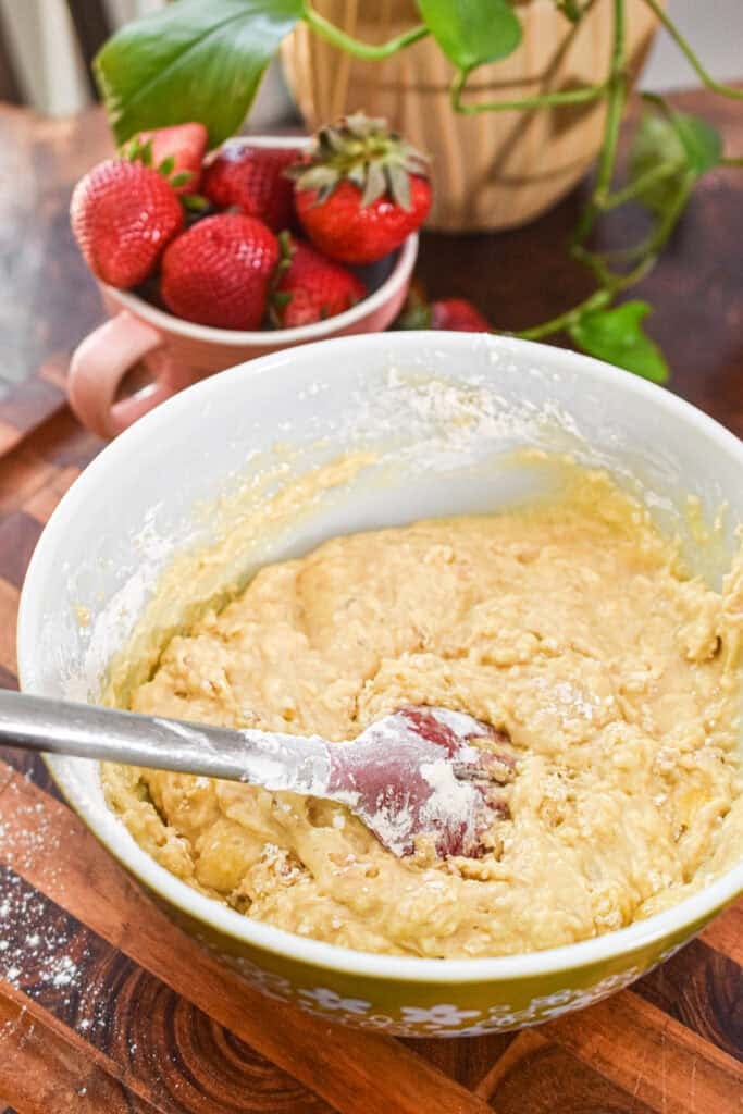 This image features a bowl of strawberry muffin batter with fresh strawberries in the background, highlighting the baking process for bakery-style 