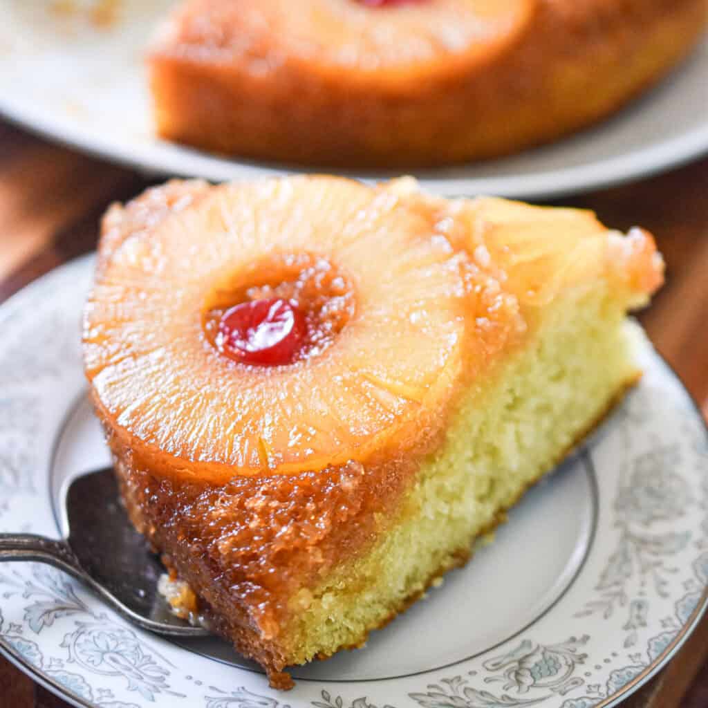 A piece of the sourdough pineapple upside down cake is on a plate with the rest of the cake behind it.