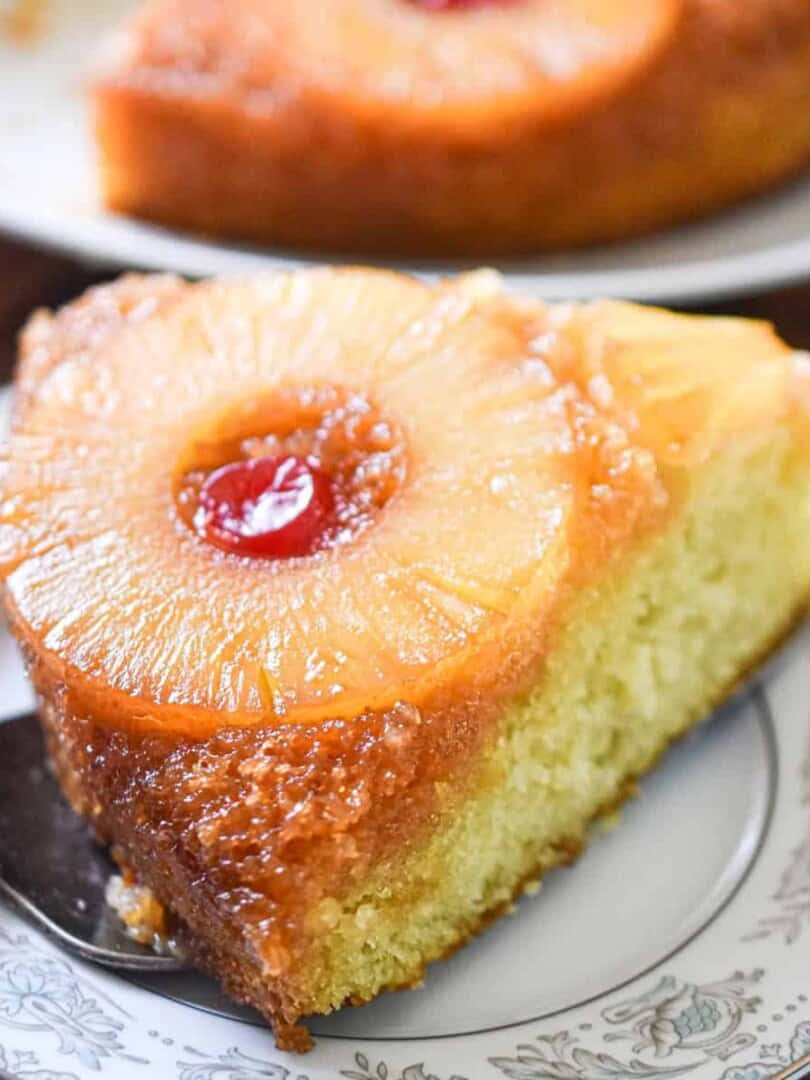 A piece of the sourdough pineapple upside down cake is on a plate with the rest of the cake behind it.