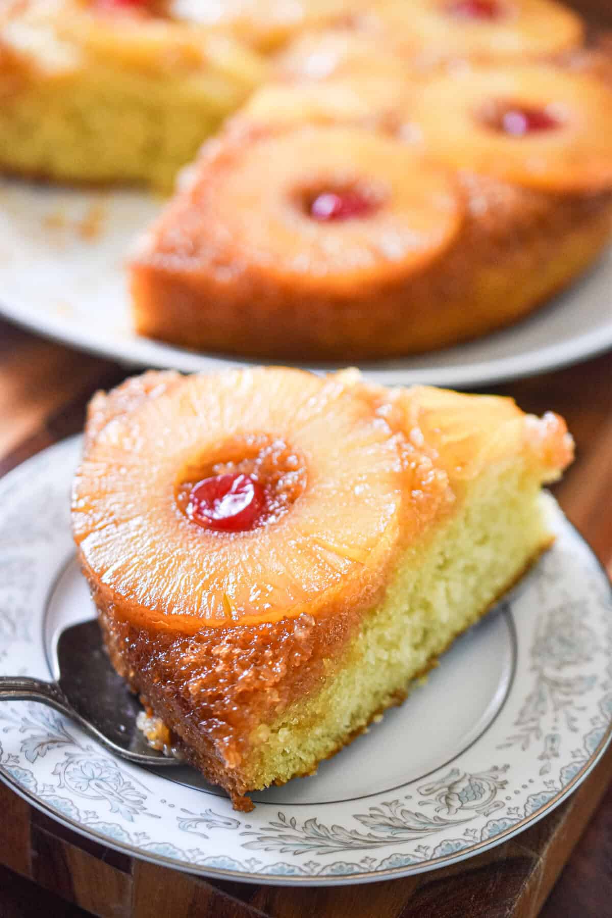 A piece of the sourdough pineapple upside down cake is on a plate with the rest of the cake behind it.