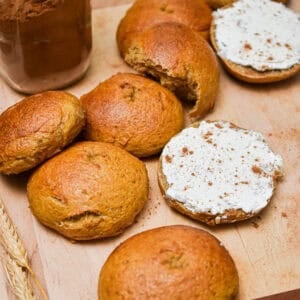 The sourdough pumpkin bagels are on a cutting board and one bagel is cut in half and shmeared with cream cheese.