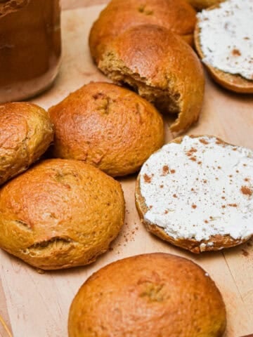 The sourdough pumpkin bagels are on a cutting board and one bagel is cut in half and shmeared with cream cheese.