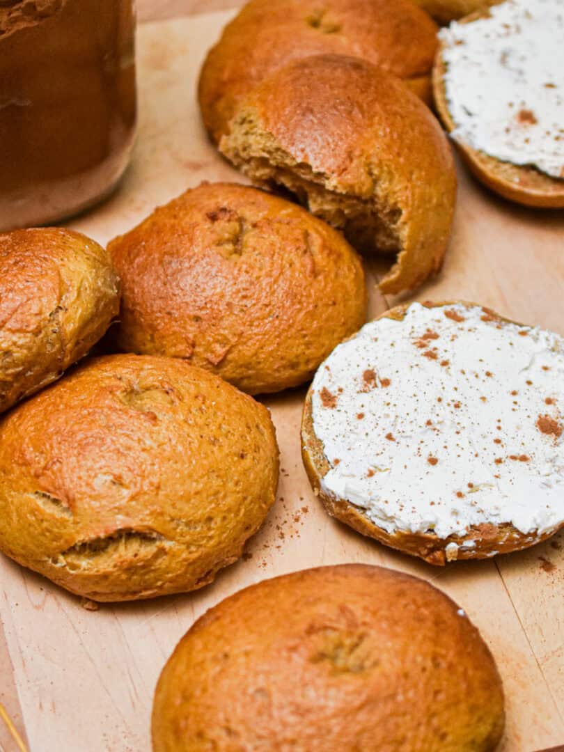 The sourdough pumpkin bagels are on a cutting board and one bagel is cut in half and shmeared with cream cheese.