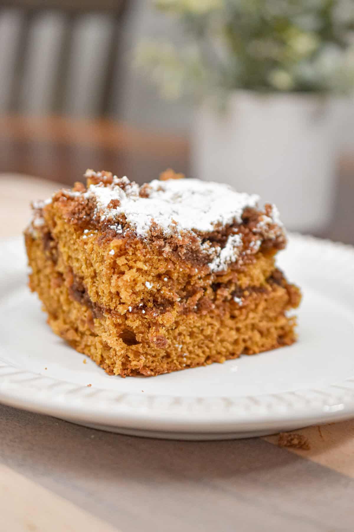The sourdough pumpkin coffee cake is on a white plate with a plant and a chair behind it.