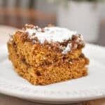 The sourdough pumpkin coffee cake is topped with powdered sugar on a white plate which is on a cutting board.