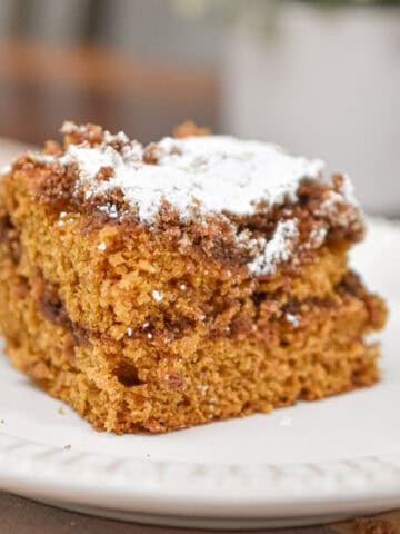 The sourdough pumpkin coffee cake is topped with powdered sugar on a white plate which is on a cutting board.